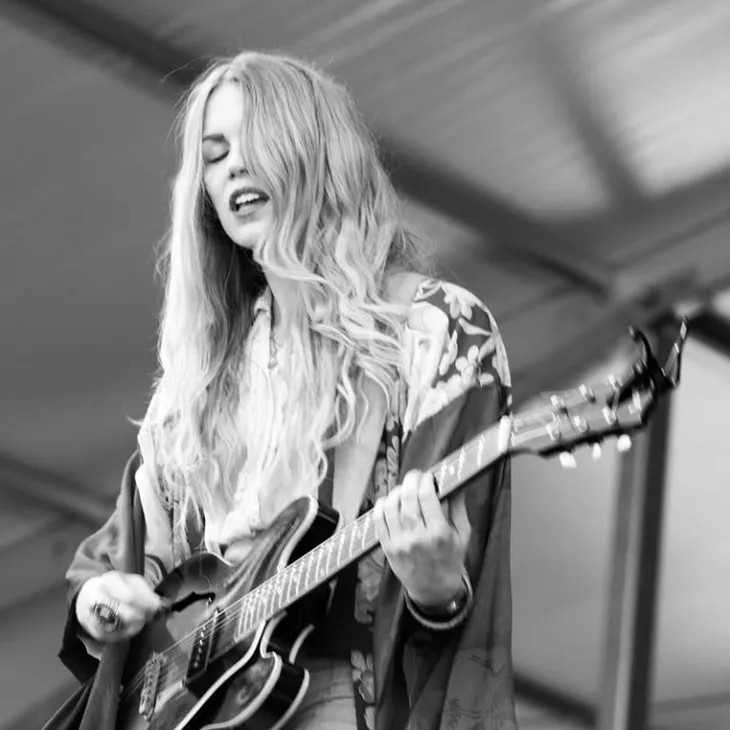 Black-and-white photo of musician Kristin Diable performing onstage, singing while playing an electric guitar.