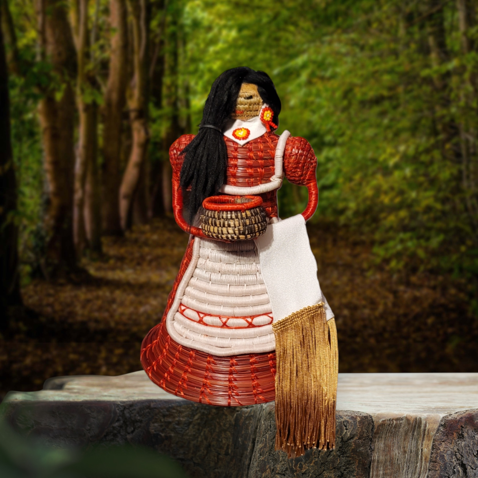 Handmade pine-needle doll by Choctaw artist Becky Meziere, posed against a wooded forest backdrop. The doll has long black hair, a woven red-and-white dress with a white apron, beaded floral earrings, and a woven basket at the waist. A long fringed woven textile hangs from the front of the figure.