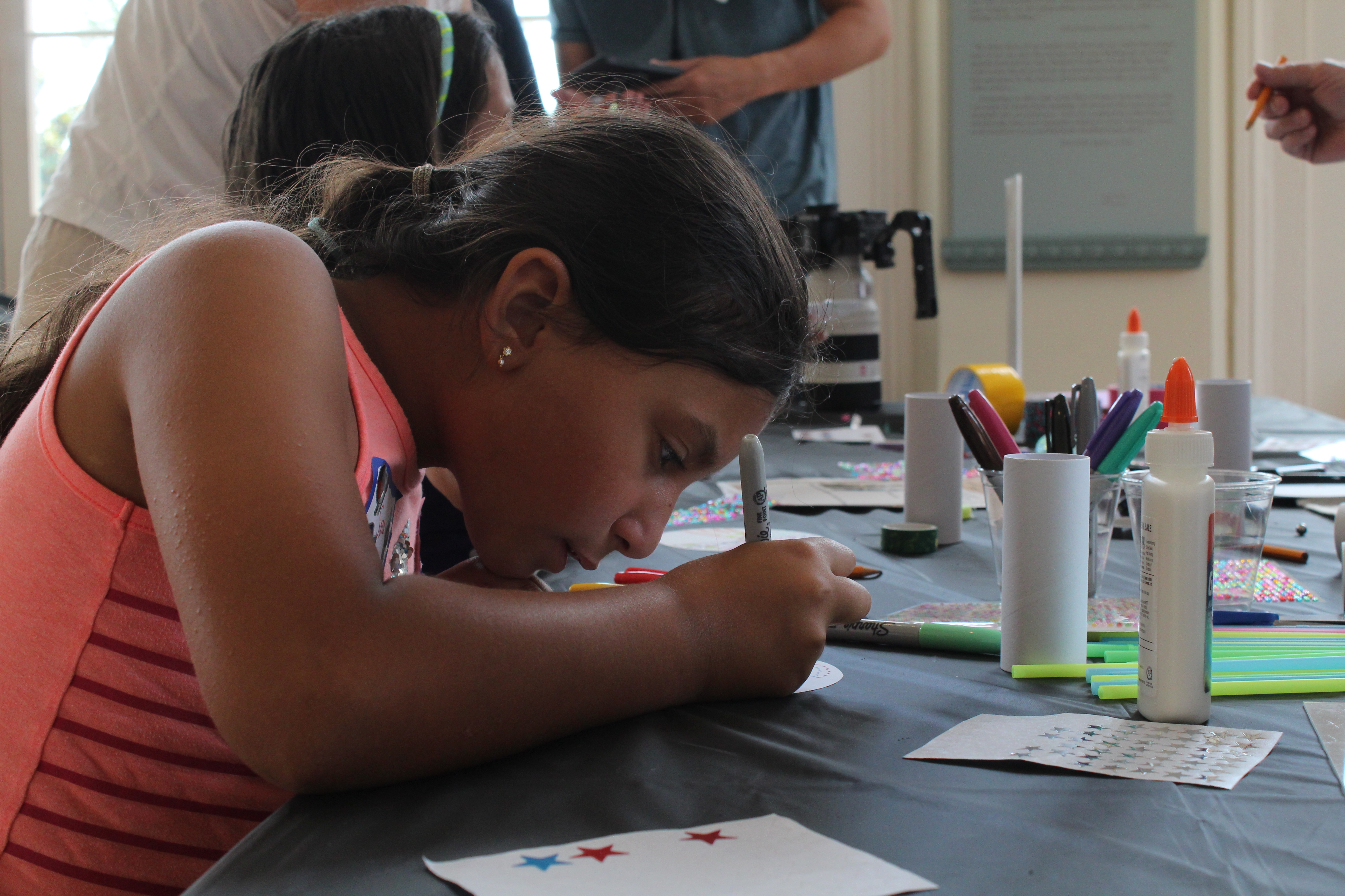 A young girl wearing a coral-colored sleeveless shirt leans over a table, concentrating on a hands-on arts and crafts project. The table is covered with colorful markers, glue, stickers, paper, and other craft supplies. Other participants and adults are visible in the background of the brightly lit room.