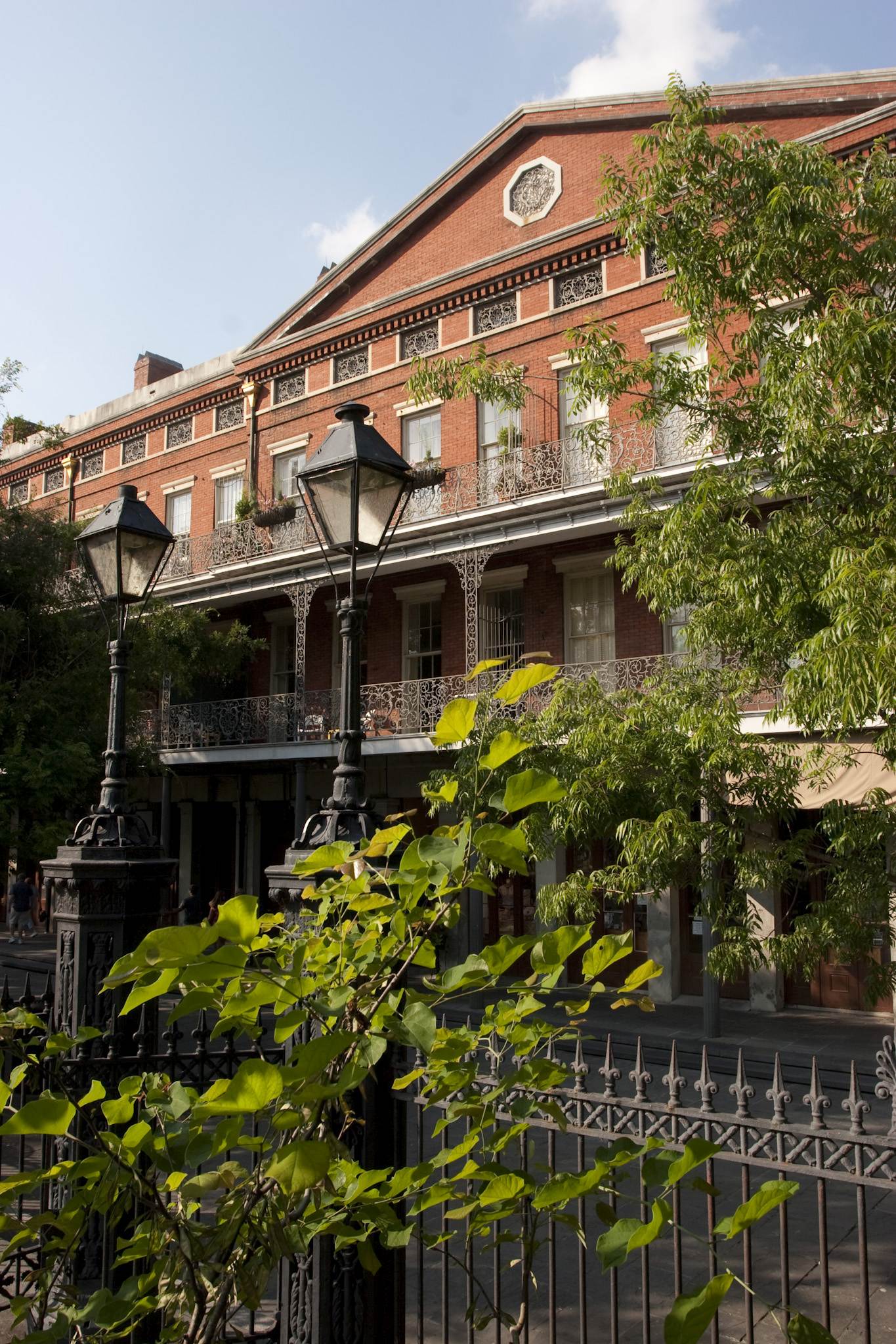 Exterior view of the 1850 House in New Orleans, a red-brick historic building with ironwork balconies and tall windows, seen from behind a wrought-iron fence. Two black streetlamps and leafy green trees frame the building.