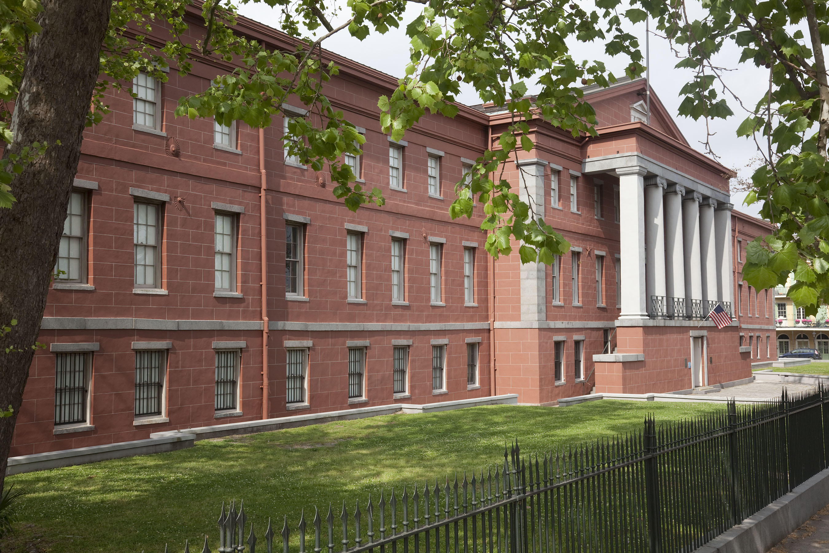 Exterior view of the New Orleans Jazz Museum at the Old U.S. Mint, a red historic building with white columns and an American flag near the entrance. Large trees frame the foreground, casting shade over the grassy lawn beside the building.