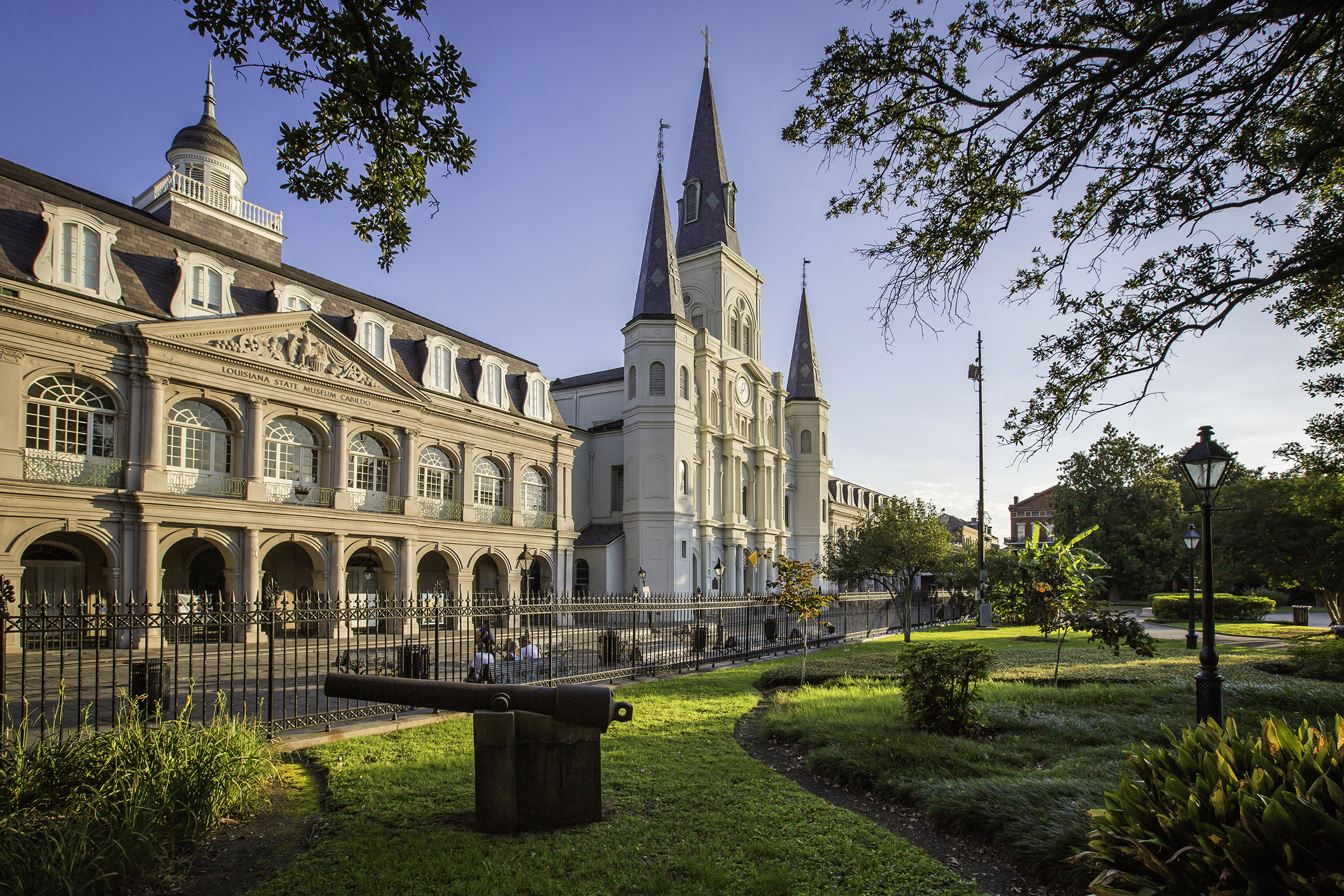View of Jackson Square in New Orleans with the Cabildo on the left and St. Louis Cathedral in the center, framed by trees and landscaped greenery. A historic cannon sits in the foreground near an iron fence, with warm sunlight illuminating the buildings.
