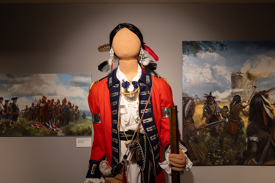 Alt text: A museum gallery display featuring a mannequin dressed in a red military-style coat with blue trim, white shirt, beaded accessories, earrings, feathers, and a long firearm. Large paintings of Revolutionary-era scenes with Indigenous figures and soldiers hang on the wall behind the mannequin.