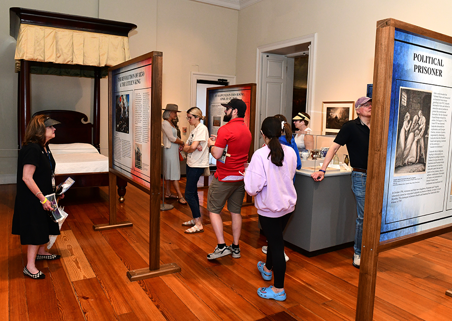 Visitors explore interpretive panels and artifacts inside the Bienvenue Lafayette exhibition at the Cabildo in New Orleans. The display includes a historic four-poster bed and information panels describing Lafayette’s life and legacy during his 1825 visit to the city.