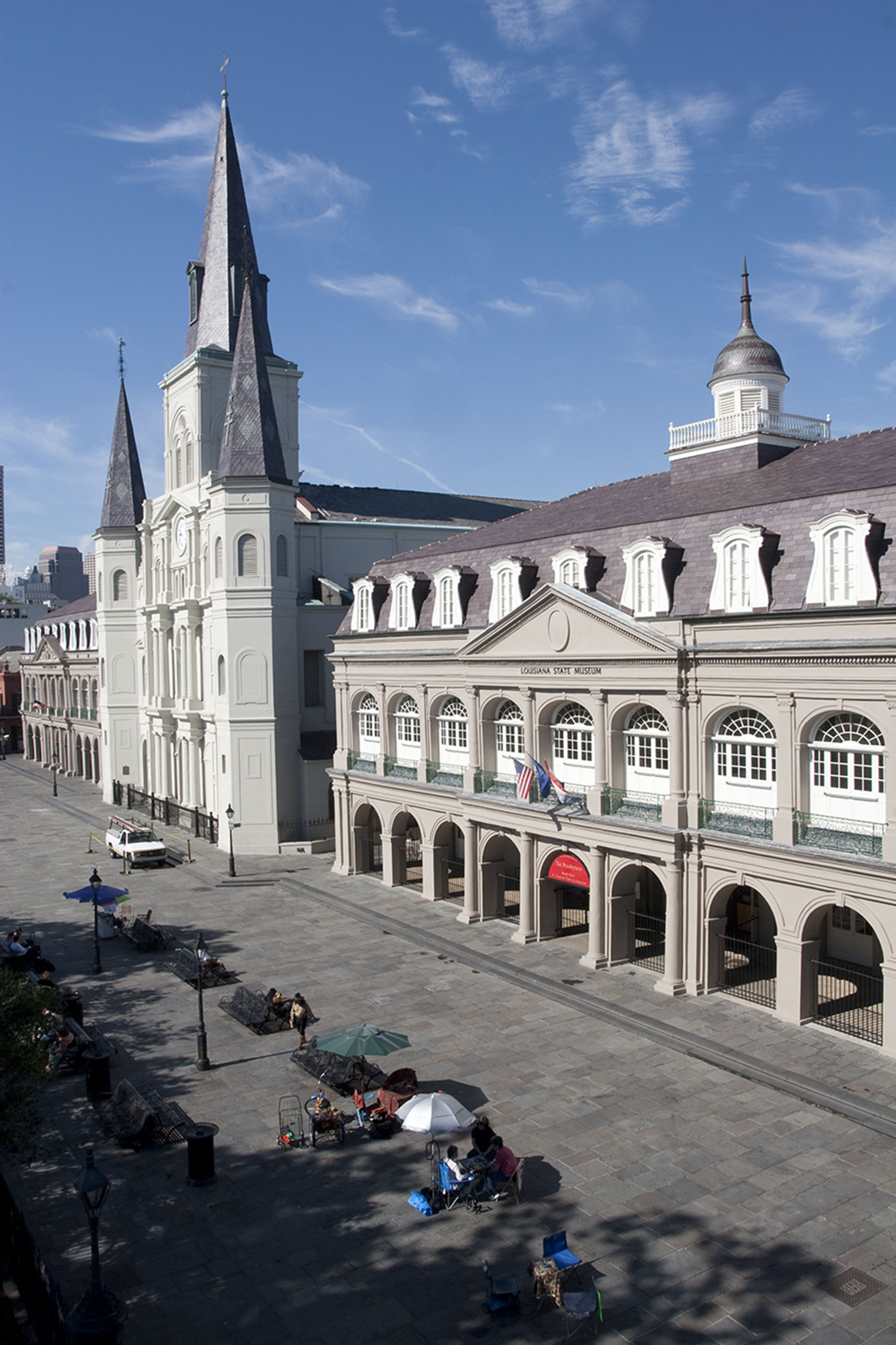 Elevated view of Jackson Square in New Orleans showing St. Louis Cathedral beside the Presbytère, a Louisiana State Museum building with arched galleries, dormer windows, and flags on the balcony. People sit and gather in the paved square below under umbrellas, with a bright blue sky overhead.