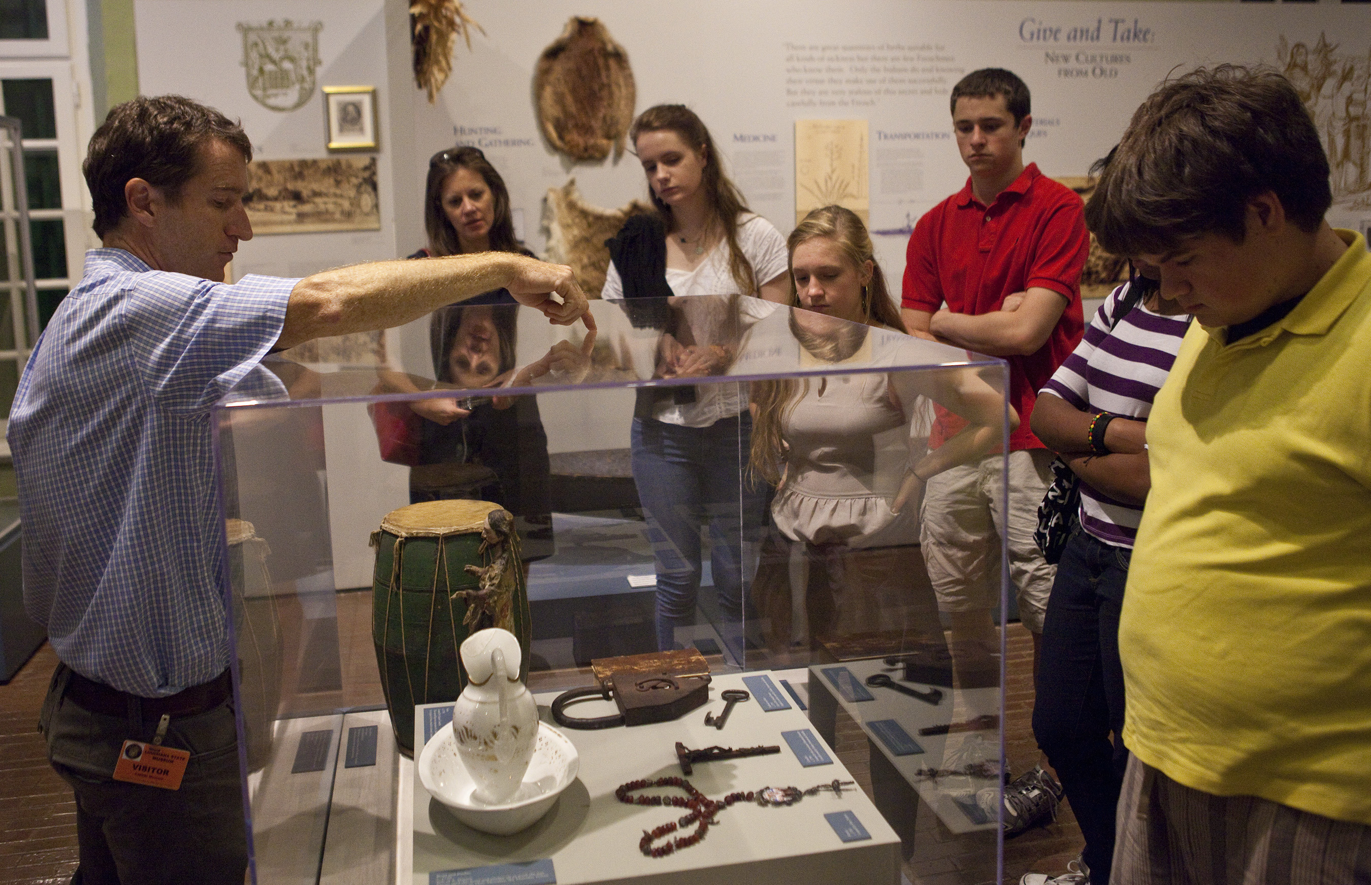 A museum curator points to objects inside a glass display case while a group of students and visitors listen attentively. The case holds historical artifacts, including a drum, a ceramic vessel, large iron keys, and a rosary. Exhibit panels and images are visible on the wall behind them.