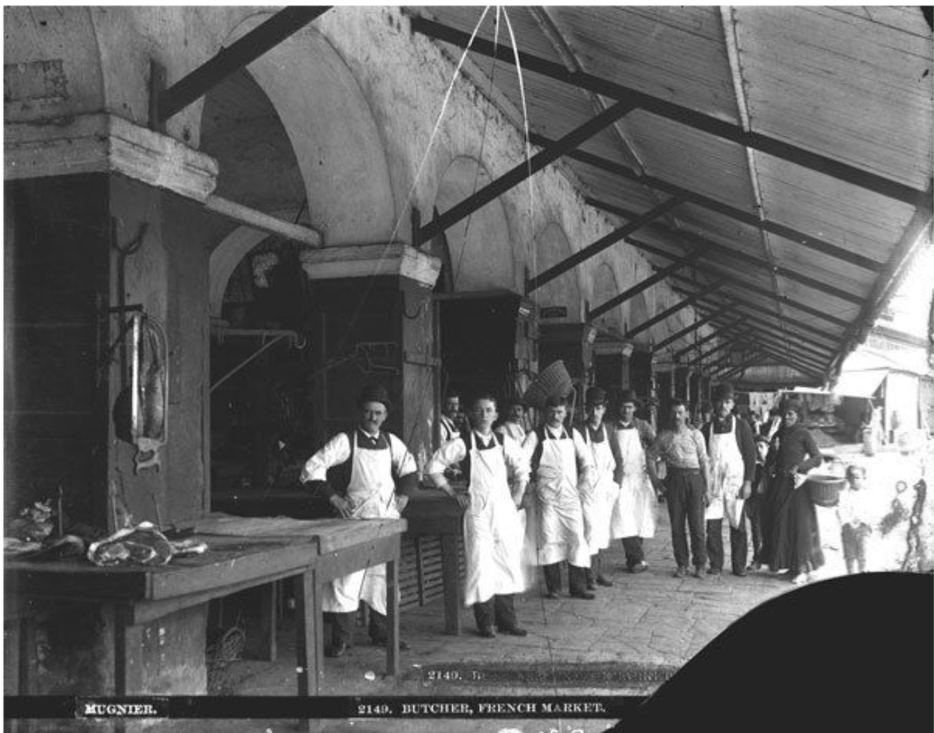 Black-and-white photograph of a row of butchers standing outside their stalls at the French Market in New Orleans. The men wear white aprons and stand beneath a covered arcade with arched openings and exposed beams. Meat hangs in the foreground, and a few women and a child stand farther down the walkway.