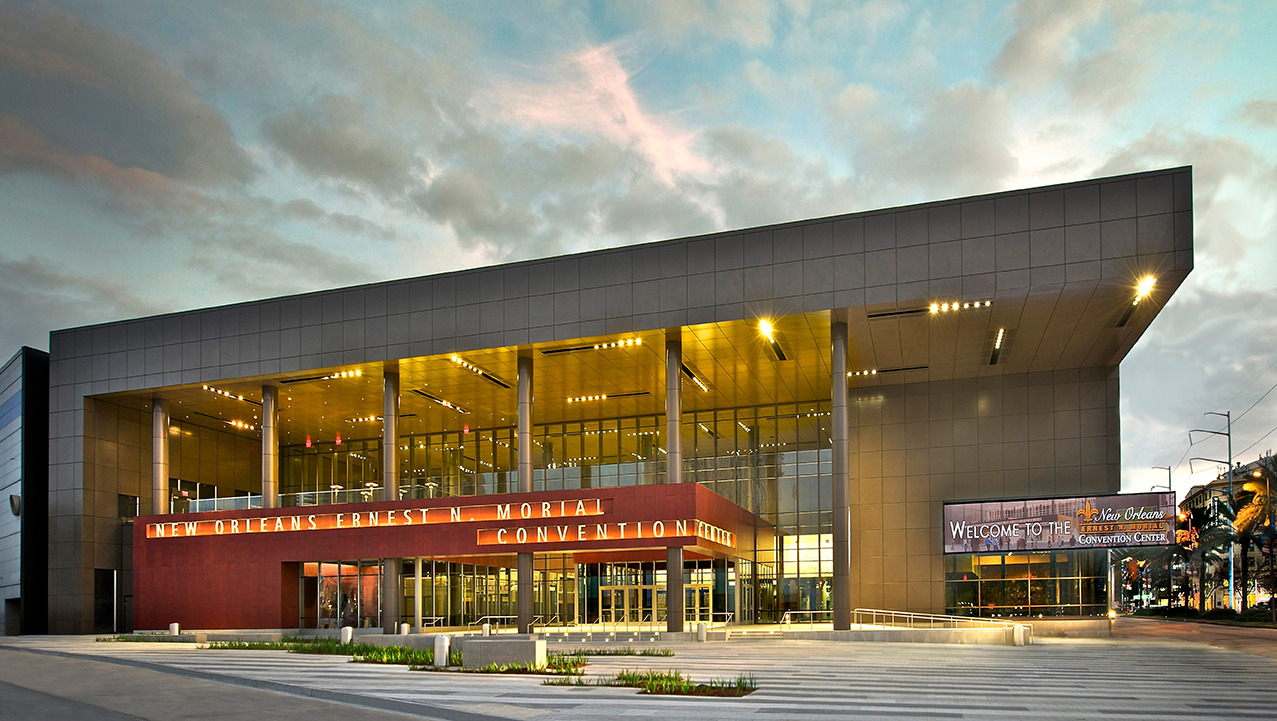 Exterior view of the Ernest N. Morial Convention Center in New Orleans at dusk. The modern glass-and-metal building is illuminated from within, with the convention center name displayed above the entrance. A large welcome sign is visible on the right side of the building, while dramatic clouds fill the evening sky overhead.