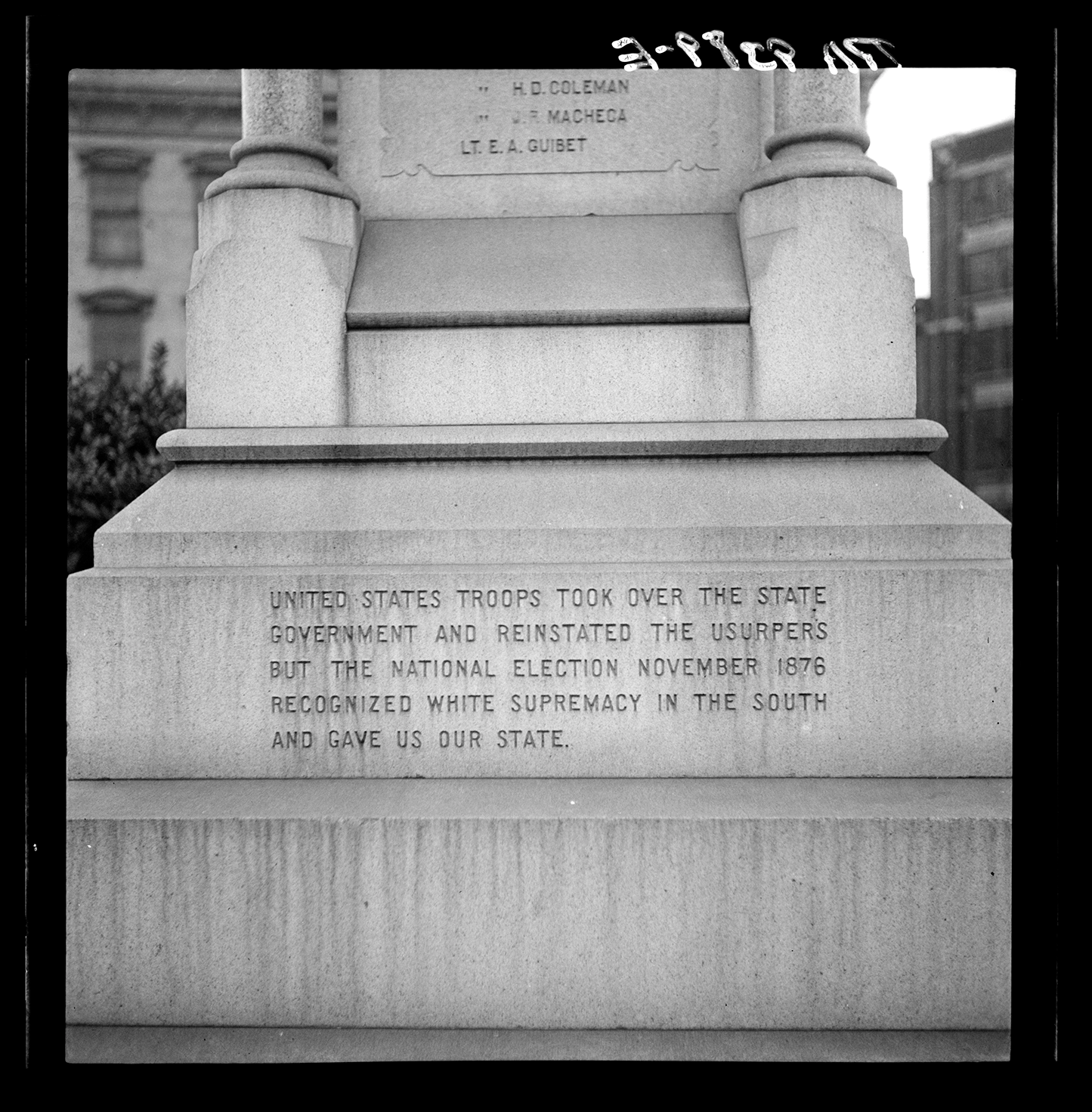 Black-and-white photograph of the base of a stone monument with an engraved inscription. The text states that U.S. troops took over the state government during Reconstruction and claims that the 1876 national election “recognized white supremacy in the South.” The carved message reflects a Lost Cause interpretation of events.