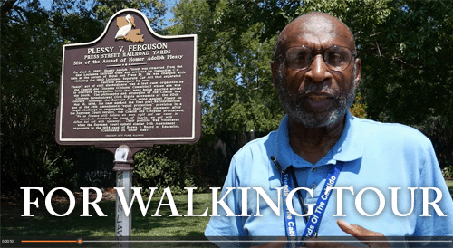A man in a blue polo shirt stands beside the Plessy v. Ferguson historical marker in New Orleans. Large white text across the bottom reads, “FOR WALKING TOUR.”