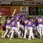 Members of the LSU Shreveport Pilots baseball team wearing purple and gold uniforms celebrate together on the field, cheering and embracing after a victory under stadium lights. Advertising signs line the outfield wall in the background.