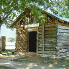 Photograph of a small rustic wooden blacksmith shop with a tin roof and a sign reading “Blacksmith” above the doorway. The log structure sits under the shade of large trees, with a brick fence and grassy yard in the background.