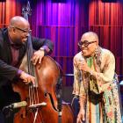 A smiling jazz vocalist and an upright bass player perform together on stage under warm purple and blue lighting. The singer holds a microphone and leans toward the bassist, both sharing a joyful moment mid-performance. A piano and drum set are visible in the background.