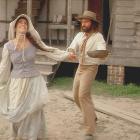 A woman in 19th-century Cajun clothing smiles and spins outdoors while a bearded man in period attire reaches toward her with a wooden bowl in his hands. They stand in front of a weathered wooden house raised on brick piers.