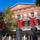 Holiday decorations in Jackson Square: Three black iron lampposts topped with bright red bows stand in front of the historic Pontalba Building, with its red brick façade, lacy wrought-iron balconies, and a clear blue sky overhead.