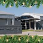 Exterior view of the Wedell-Williams Aviation & Cypress Sawmill Museum in Patterson, Louisiana, on a sunny day. The modern gray building features a curved entrance canopy, tall windows, and an American flag on a pole to the right. The image is framed at the top and bottom with illustrated green pine branches and glowing holiday string lights.