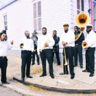 Sporty’s Brass Band poses outdoors on a New Orleans street corner, wearing black-and-white attire and holding brass instruments including trumpets, trombones, and a sousaphone, with colorful historic buildings in the background.