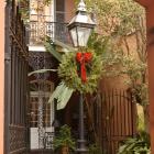 Photograph of a traditional New Orleans Creole courtyard framed by a stucco archway. At the center stands a black iron gas-style lamppost decorated with a green holiday wreath and red bow. Wrought-iron gates, brick paving, potted plants, and a two-story building with decorative iron balconies and shuttered doors surround the intimate courtyard space.