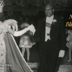 Black-and-white photograph of a formal Black Carnival ball. A young woman wearing an ornate crown, embellished gown, long gloves, and holding a decorative scepter is escorted by an older man in a tuxedo and white gloves as they walk across a ballroom floor. Dancers in matching costumes stand behind them beneath draped curtains. Overlaid gold text reads, “Origins of New Orleans Black Carnival Society: The Story of the Illinois Clubs,” with The Presbytère logo. A caption at bottom left identifies the scene as