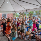 A large crowd of people dances with their hands raised under a white event tent during an outdoor festival. Attendees of various ages fill the space, some standing and some seated in folding chairs, with bright clothing and expressive movement. Behind the tent, trees, vendor tents, and the exterior of a peach-colored building are visible.