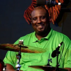 A smiling Black man wearing a bright green shirt sits at a drum set. He is positioned behind cymbals and microphones on a dimly lit stage, with orange and black stage decorations visible in the background.