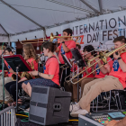 A large student jazz ensemble performs onstage under a white festival tent. Musicians in matching bright coral shirts play trombones, saxophones, and trumpets while reading from music stands. A banner behind them reads “International Jazz Day Festival.” A sound engineer operates a mixing console in the foreground.