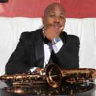 A handsome Black man in a black tuxedo sits on a white leather bench, resting his chin on his hand in a thoughtful pose. A polished brass saxophone lies on the table in front of him, reflecting light. A red patterned wall forms the background.