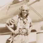 A smiling woman in a flight suit stands in front of an airplane, wearing a parachute harness and aviator gear.
