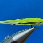 Close-up of a bright chartreuse and white fishing fly held in a metal fly-tying vise against a blue background. The fly has a red head, silver eye detail, black accent lines, and a hook extending beneath the body.