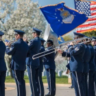U.S. Air Force Band members in dress uniforms perform outdoors with brass instruments and drums. American and Air Force flags wave above the band, with rows of white headstones visible in the background.