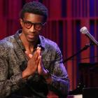 Kyle Roussel sits at a piano onstage beside a microphone, hands clasped, with warm red and purple lighting in the background.