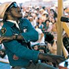 Professor Longhair performs at a piano on an outdoor stage, wearing sunglasses and a cap, with a large festival crowd in the background.