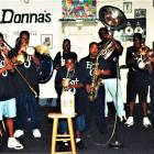 A group of seven musicians, including children, perform together indoors, playing brass and percussion instruments. A large “Donna’s” sign and framed artwork are visible on the wall behind them.  