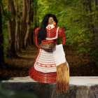 Handmade pine-needle doll by Choctaw artist Becky Meziere, posed against a wooded forest backdrop. The doll has long black hair, a woven red-and-white dress with a white apron, beaded floral earrings, and a woven basket at the waist. A long fringed woven textile hangs from the front of the figure.