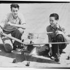 Black-and-white photograph of two young men crouching outdoors beside several handmade model airplanes, including a large glider and smaller propeller-driven aircraft.