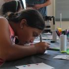 A young girl wearing a coral-colored sleeveless shirt leans over a table, concentrating on a hands-on arts and crafts project. The table is covered with colorful markers, glue, stickers, paper, and other craft supplies. Other participants and adults are visible in the background of the brightly lit room.