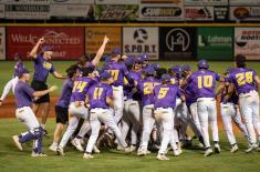 Members of the LSU Shreveport Pilots baseball team wearing purple and gold uniforms celebrate together on the field, cheering and embracing after a victory under stadium lights. Advertising signs line the outfield wall in the background.