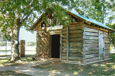 Photograph of a small rustic wooden blacksmith shop with a tin roof and a sign reading “Blacksmith” above the doorway. The log structure sits under the shade of large trees, with a brick fence and grassy yard in the background.