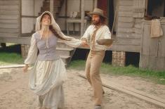 A woman in 19th-century Cajun clothing smiles and spins outdoors while a bearded man in period attire reaches toward her with a wooden bowl in his hands. They stand in front of a weathered wooden house raised on brick piers.