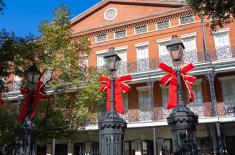 Holiday decorations in Jackson Square: Three black iron lampposts topped with bright red bows stand in front of the historic Pontalba Building, with its red brick façade, lacy wrought-iron balconies, and a clear blue sky overhead.