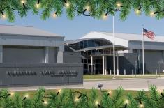 Exterior view of the Wedell-Williams Aviation & Cypress Sawmill Museum in Patterson, Louisiana, on a sunny day. The modern gray building features a curved entrance canopy, tall windows, and an American flag on a pole to the right. The image is framed at the top and bottom with illustrated green pine branches and glowing holiday string lights.