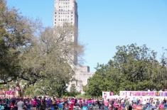 Crowds gathered outdoors during the Spanish Town Parade in Baton Rouge, with spectators wearing colorful clothing and costumes seated in folding chairs and standing along the route, trees lining the scene, and the Louisiana State Capitol tower rising prominently in the background under a clear blue sky.