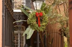 Photograph of a traditional New Orleans Creole courtyard framed by a stucco archway. At the center stands a black iron gas-style lamppost decorated with a green holiday wreath and red bow. Wrought-iron gates, brick paving, potted plants, and a two-story building with decorative iron balconies and shuttered doors surround the intimate courtyard space.