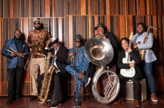 The Dirty Dozen Brass Band poses for a group portrait in a recording studio, with members standing and seated against a wood-paneled wall while holding brass instruments including trombone, saxophones, trumpet, sousaphone, and guitar.