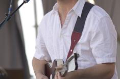Alex McMurray performs onstage, singing into a microphone while playing an electric guitar. He wears a white short-sleeve shirt and sunglasses, with a guitar strap over his shoulder, during an outdoor live music performance.