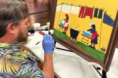 Conservator wearing blue gloves carefully cleans a framed folk-style painting on an easel, using a small brush while seated at a worktable inside a museum gallery.