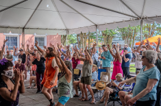 A large crowd of people dances with their hands raised under a white event tent during an outdoor festival. Attendees of various ages fill the space, some standing and some seated in folding chairs, with bright clothing and expressive movement. Behind the tent, trees, vendor tents, and the exterior of a peach-colored building are visible.