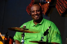 A smiling Black man wearing a bright green shirt sits at a drum set. He is positioned behind cymbals and microphones on a dimly lit stage, with orange and black stage decorations visible in the background.