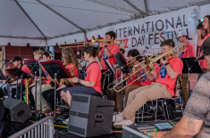 A large student jazz ensemble performs onstage under a white festival tent. Musicians in matching bright coral shirts play trombones, saxophones, and trumpets while reading from music stands. A banner behind them reads “International Jazz Day Festival.” A sound engineer operates a mixing console in the foreground.