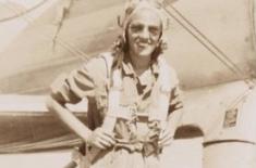A smiling woman in a flight suit stands in front of an airplane, wearing a parachute harness and aviator gear.
